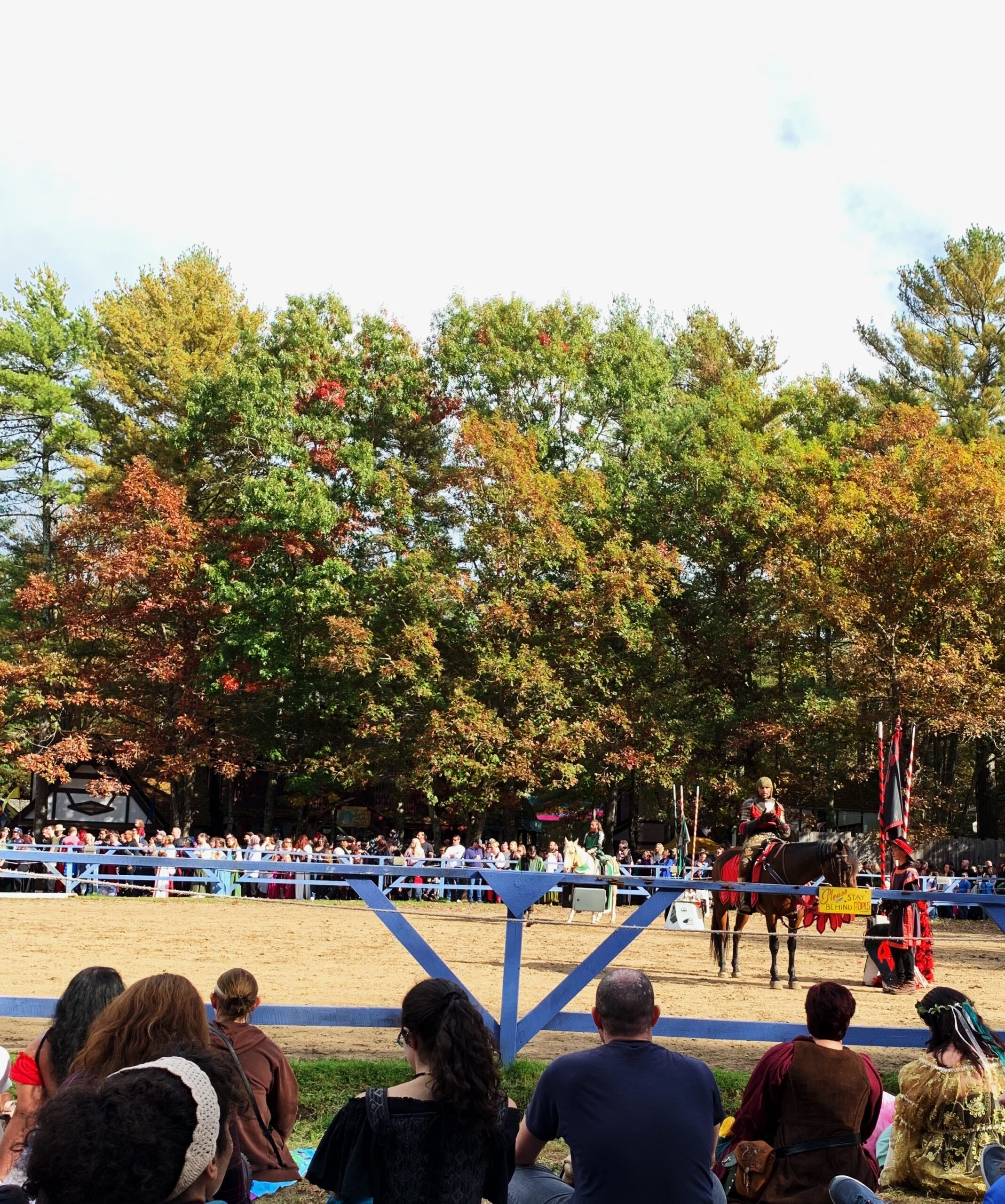 Photograph taken from the perspective of an audience member at the Tournament of King Richard's Faire, in the foreground you see other audience members, then over a blue fence is a knight dressed in red and black on horseback and behind him is another fence, more audience members and behind them are autumn trees just beginning to change color