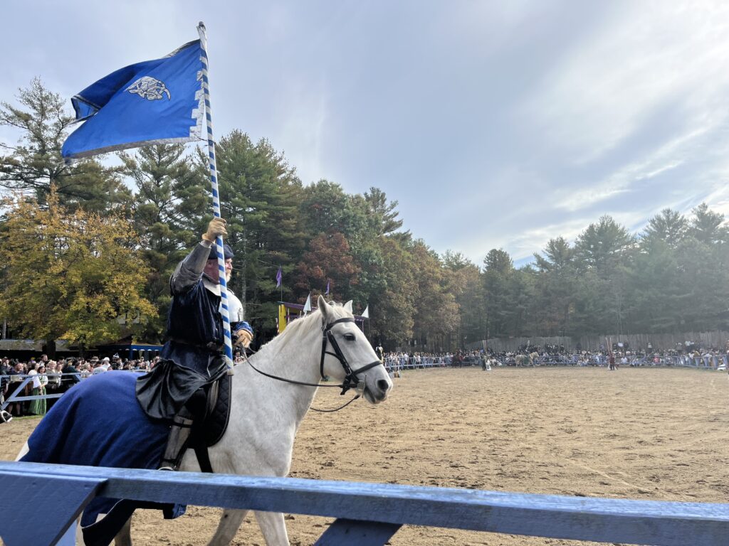 A knight in armor rides upon a white horse, holding a blue flag.